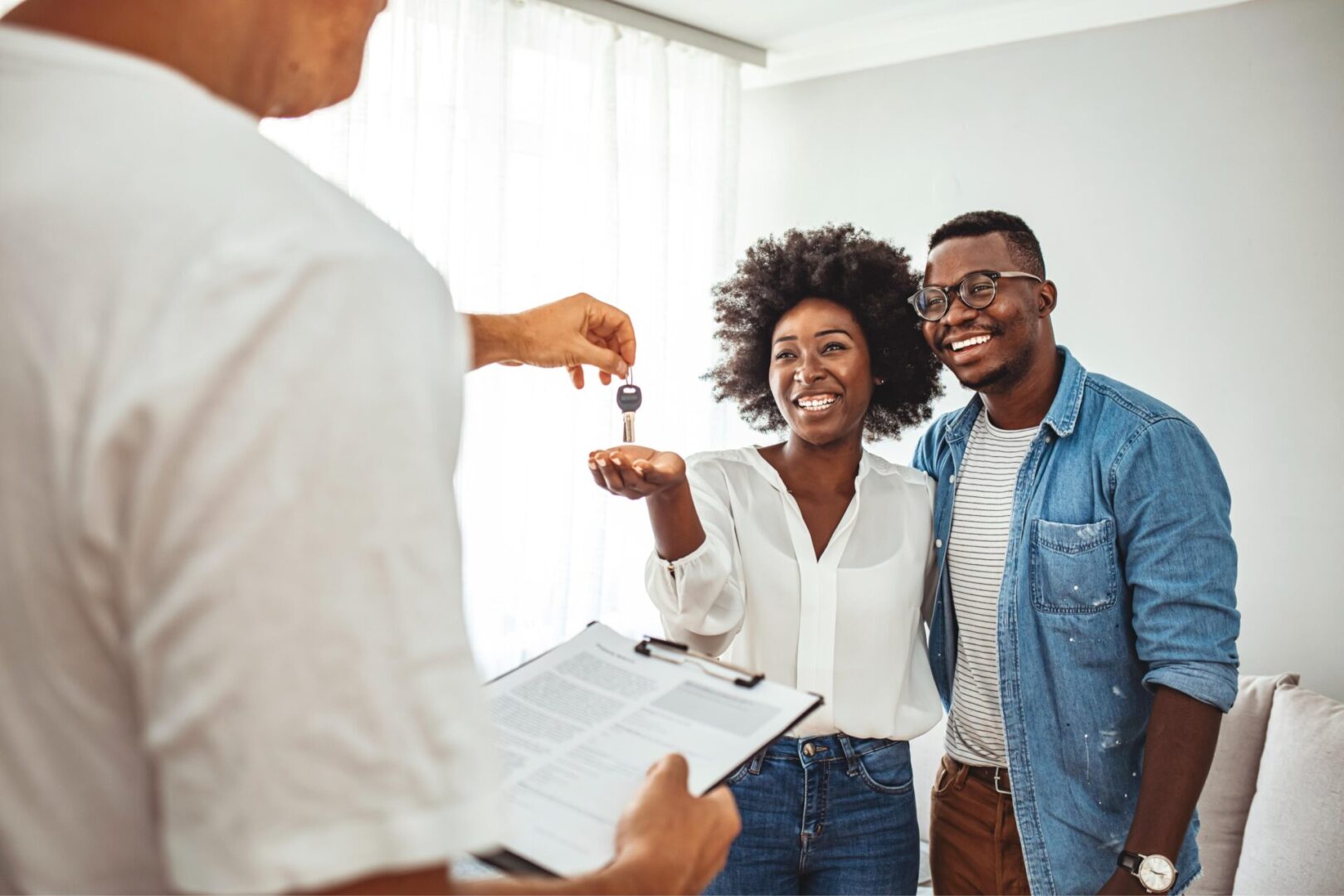 A couple receives a key from a person holding a clipboard. They appear happy and are standing in a well-lit room.