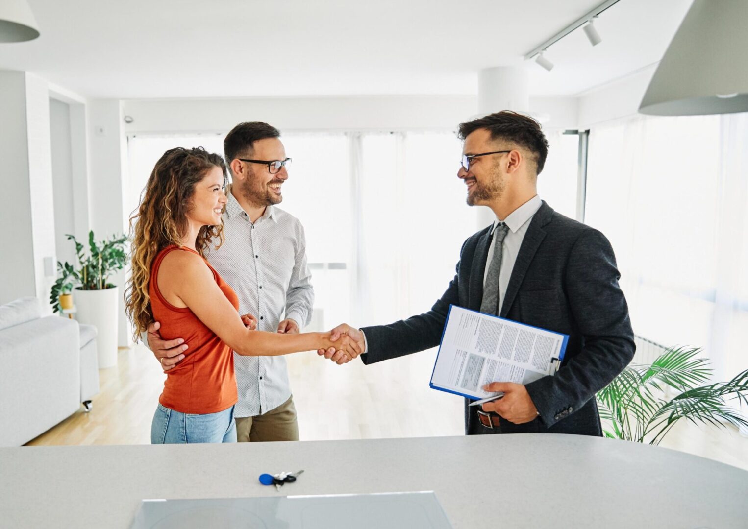 A couple shakes hands with a man in a suit holding documents in a bright room, with keys placed on the table.