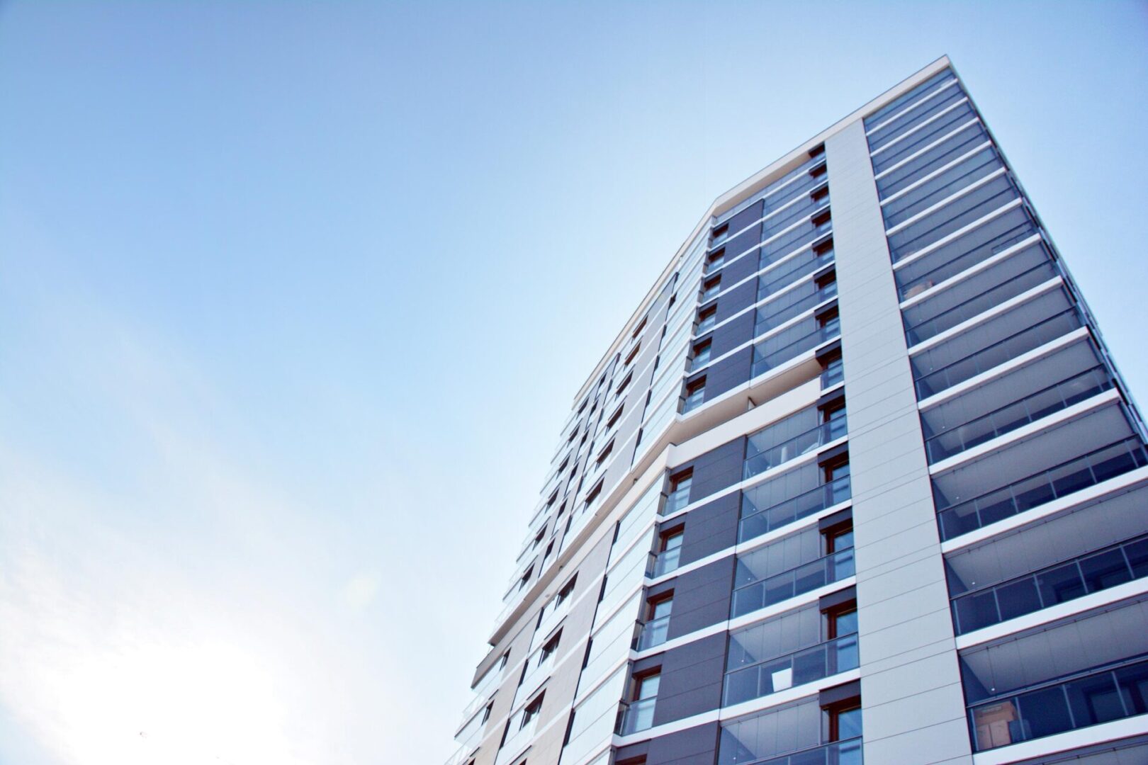 Upward view of a modern high-rise building with glass windows and balconies against a clear blue sky.