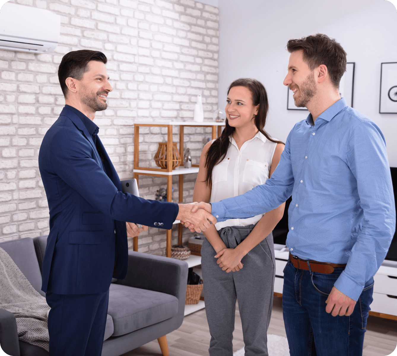 Three people in a living room; two men are shaking hands while a woman stands beside them, smiling.