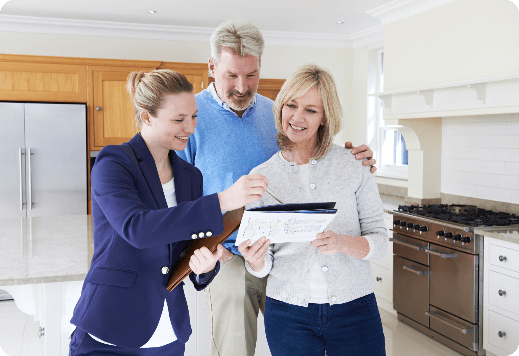 Three people in a kitchen looking at floor plans; a woman in a blue suit is showing the plans to a couple.