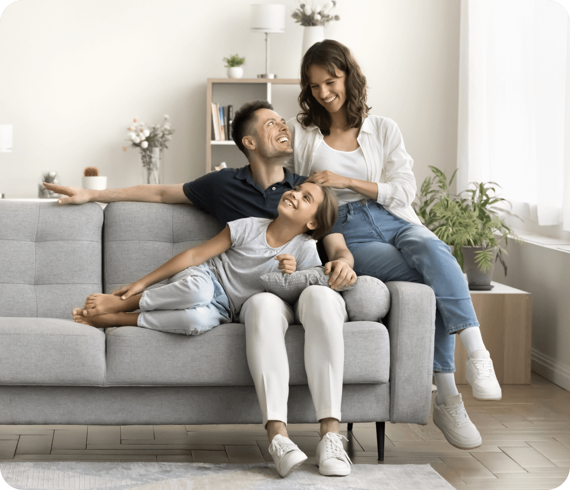 A family of three sits on a gray couch in a living room. The parents smile at each other while the child leans on the father's lap. Plants and a bookshelf are visible in the background.